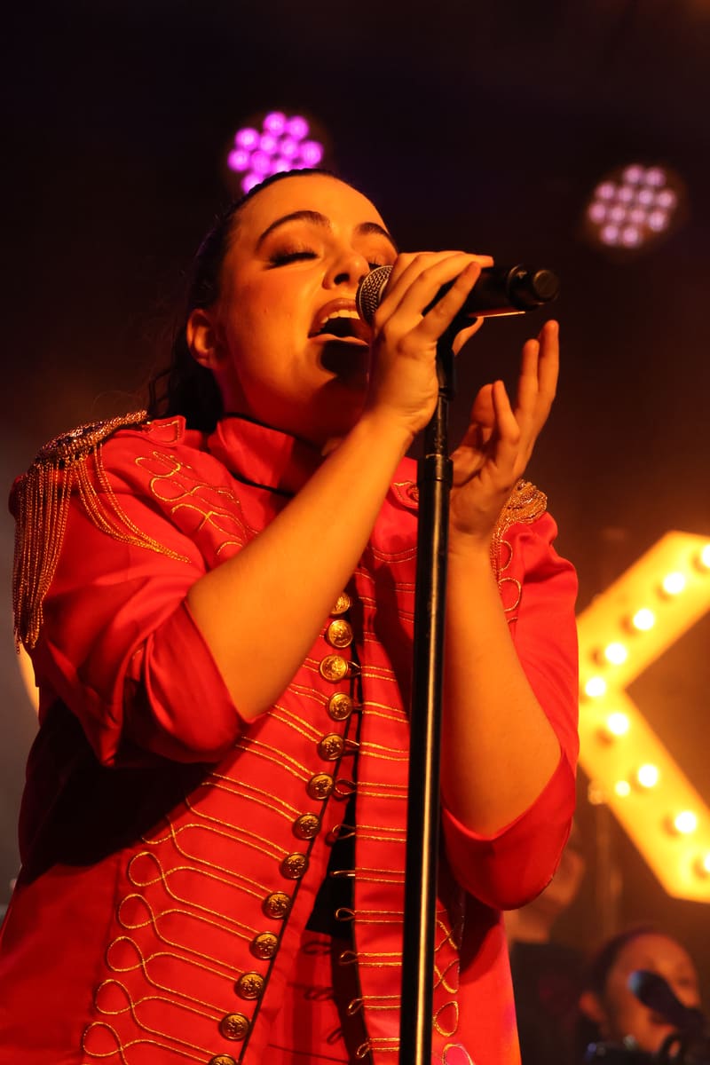 Performer in a red sequinned jacket sings into a microphone under warm stage lighting with coloured lights in the background.