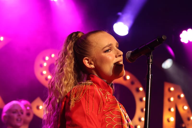 Performer singing into microphone under vibrant magenta and gold stage lighting.