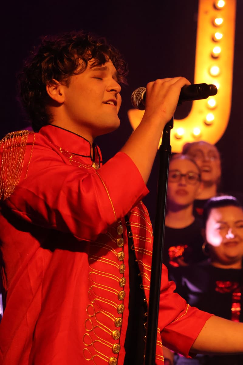Performer in red sequinned jacket singing into microphone under warm stage lighting, audience visible behind.