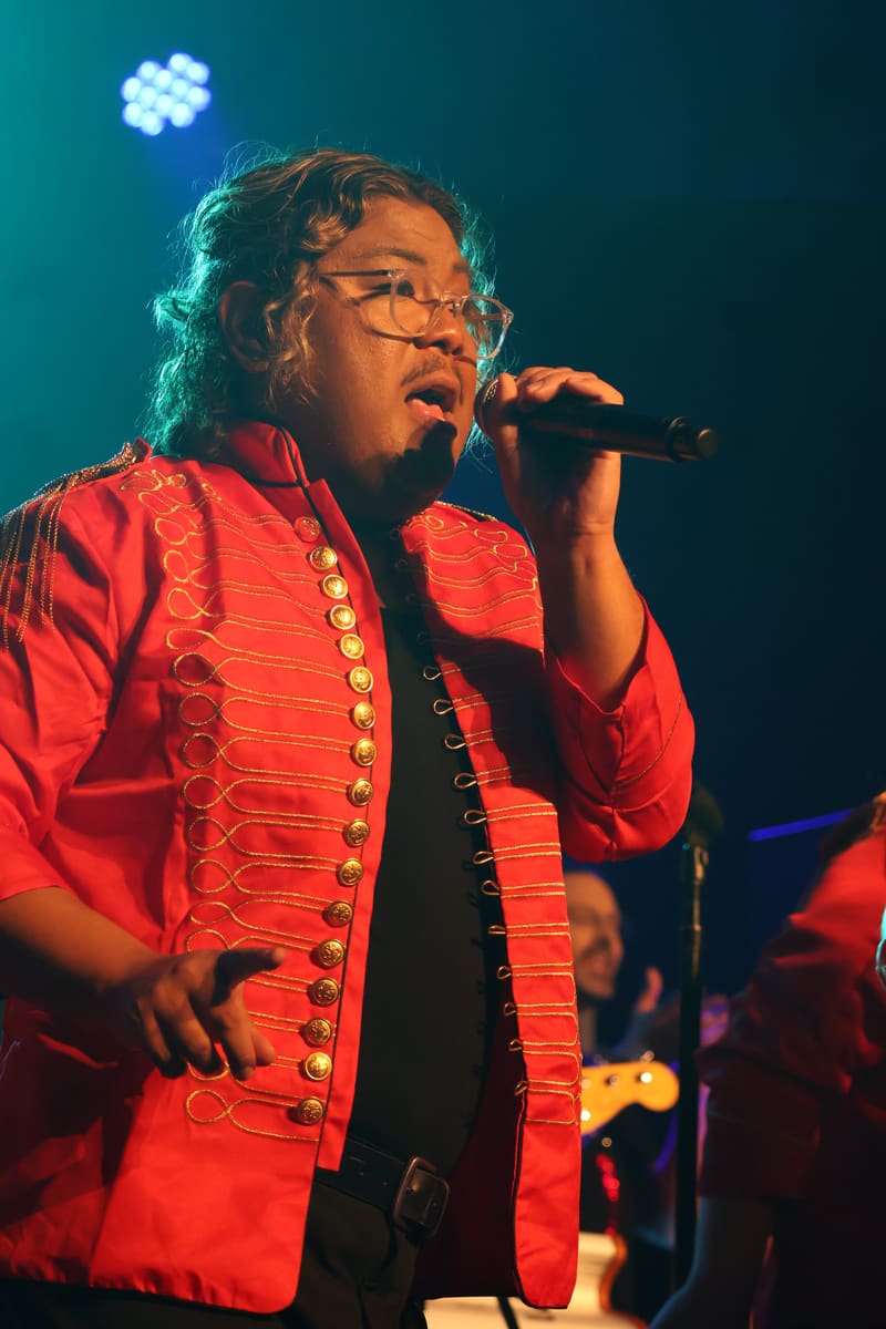 Performer in a red studded jacket singing into a microphone under teal stage lighting.