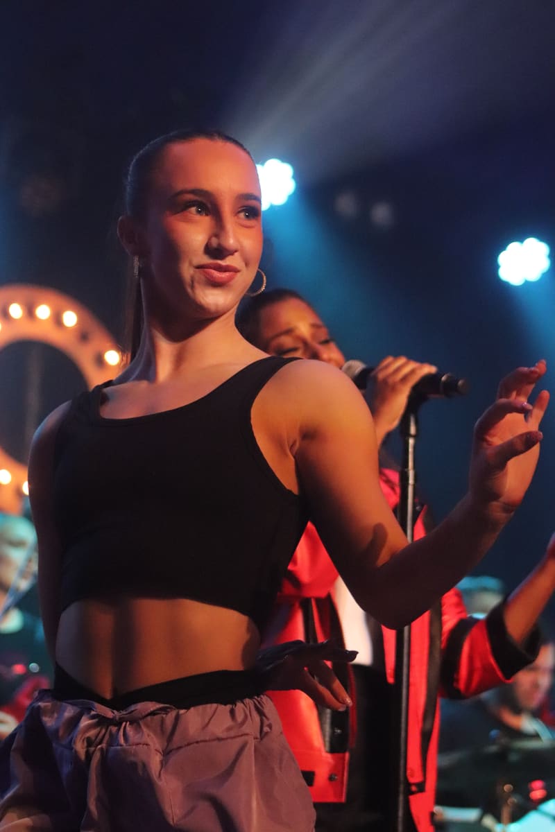 Performer on stage in black crop top and light-coloured pants, holding microphone under warm and cool stage lighting.