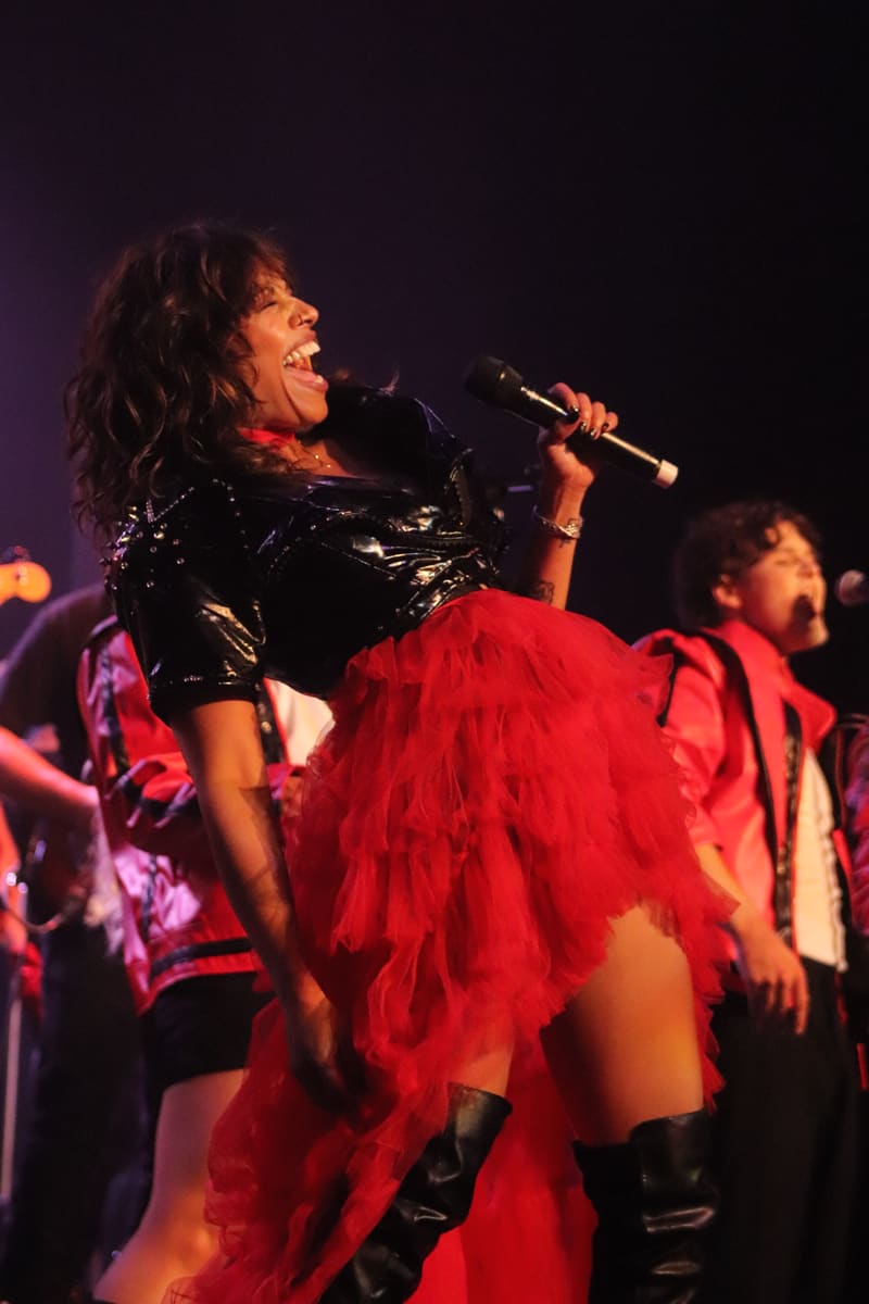Performer in red tulle skirt and black studded top singing into microphone under warm stage lighting.