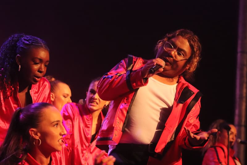 Performers in red and pink jackets dancing and singing under stage spotlights during a theatrical production.