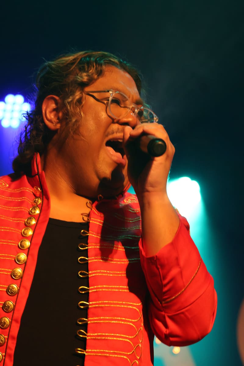 Performer singing into microphone wearing red sequined jacket and glasses under blue and white stage lighting.