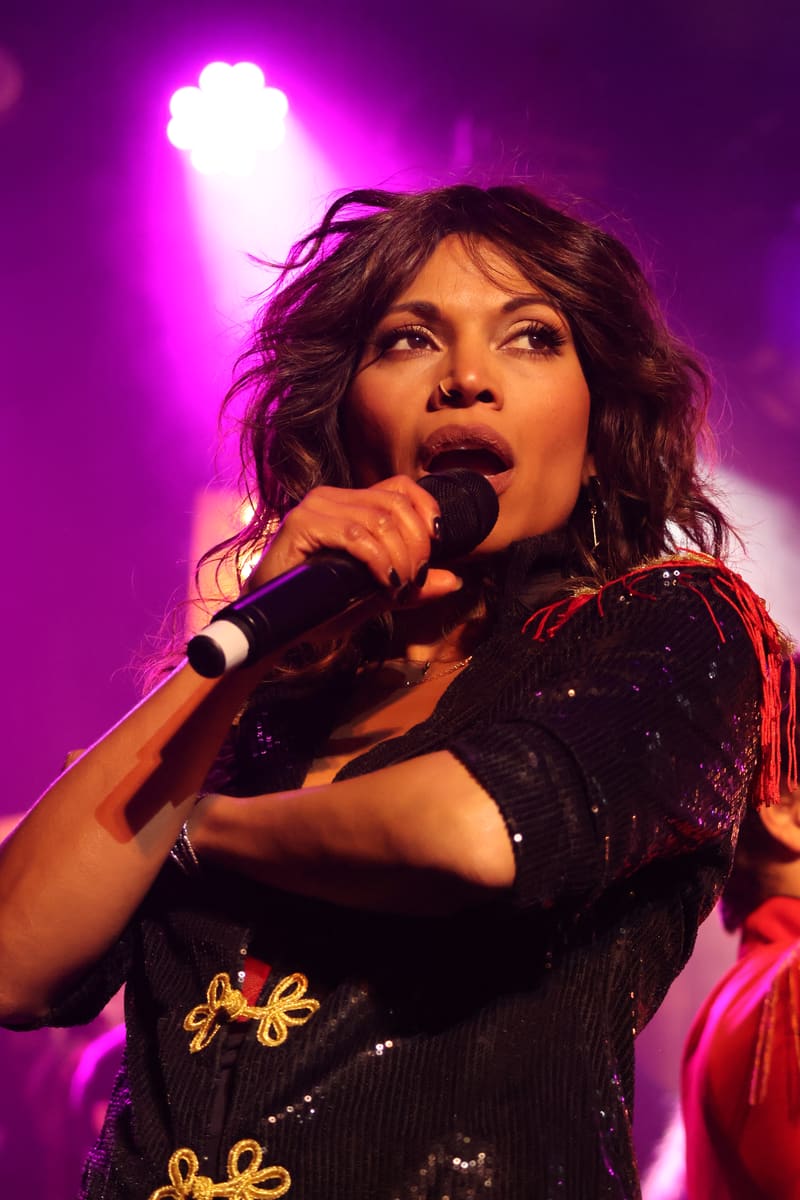 Performer singing into a microphone on stage, bathed in magenta spotlighting, wearing a sequinned black outfit.