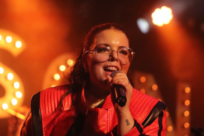 Performer in red pinstriped shirt and glasses singing into microphone beneath warm stage lighting.