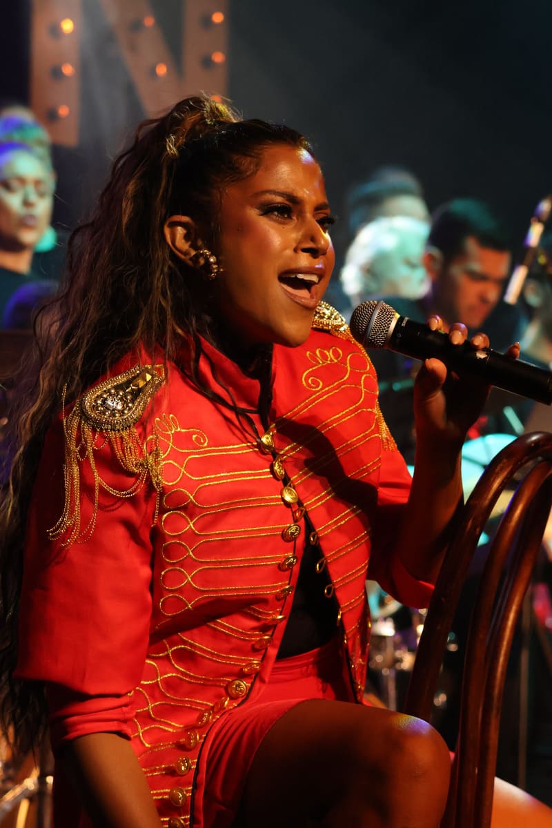 Performer in a red sequined jacket sings into a microphone under warm stage lighting.