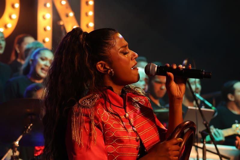 Performer singing into microphone wearing red sequinned shirt, stage-lit against backdrop with glowing signage.