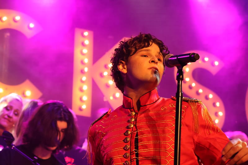 Performer in red sequinned jacket singing at microphone under magenta stage lights with illuminated backdrop.