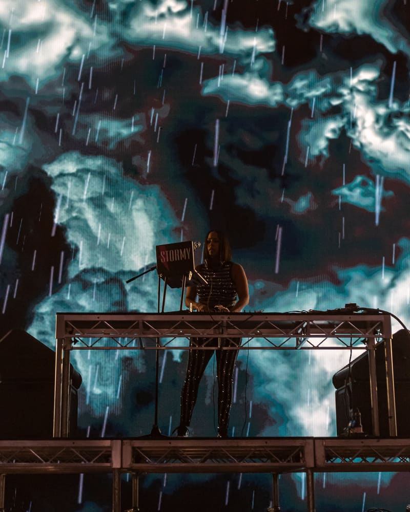 DJ silhouetted on an elevated stage rig against a dramatic projection of stormy skies and rain.