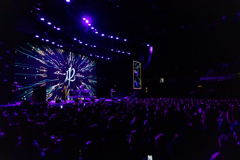Illuminated stage with dynamic purple and white projections above a crowd during a live music performance.