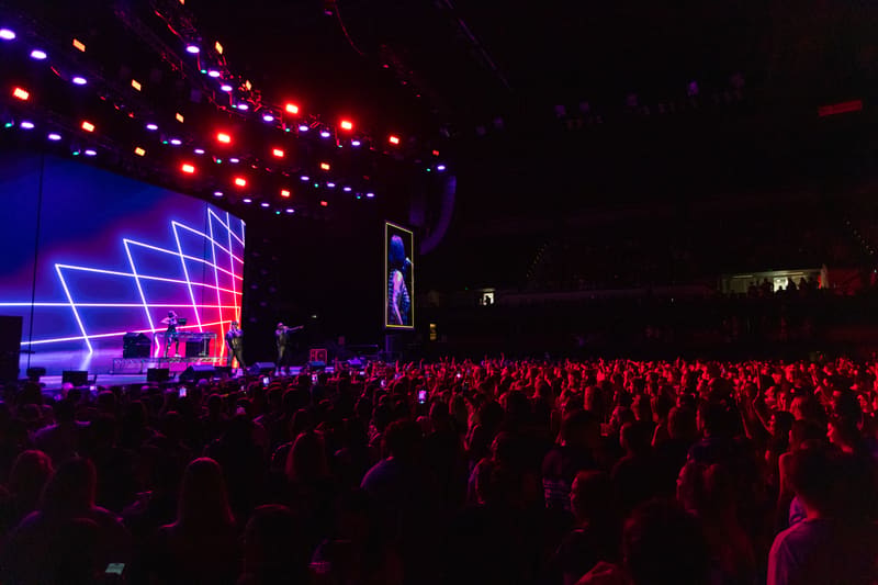 Large crowd of concert-goers facing a stage with neon blue and pink geometric screens and overhead lighting at night.