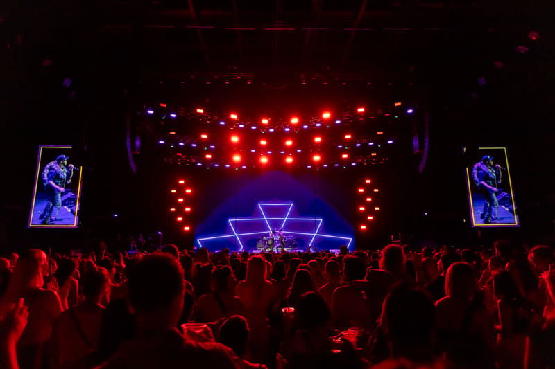 Crowd silhouetted in warm red light facing an illuminated stage with neon geometric set design and overhead lighting rig.