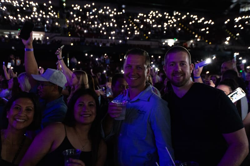 Crowd of smiling attendees bathed in purple stage lighting beneath a canopy of twinkling overhead lights.