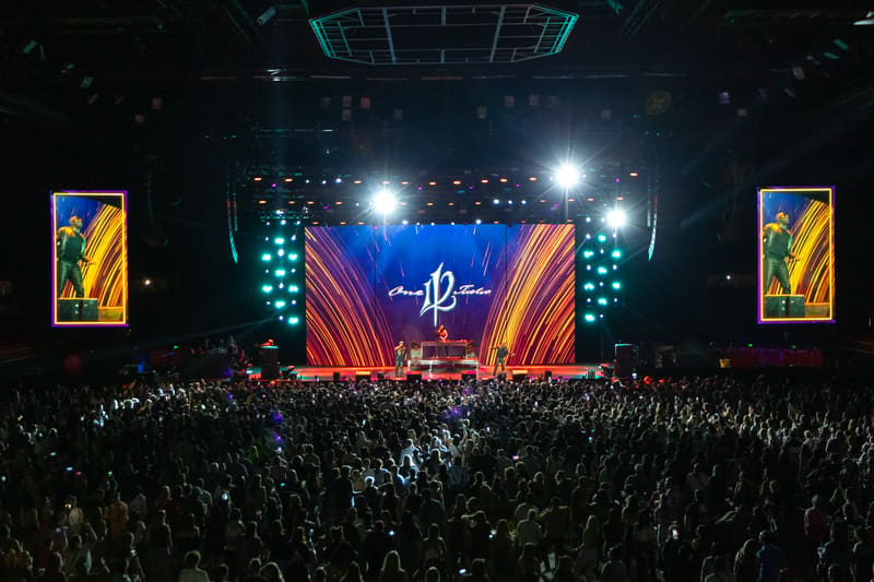 Crowded concert venue with illuminated stage featuring red and blue neon arches and central logo, audience with raised hands.