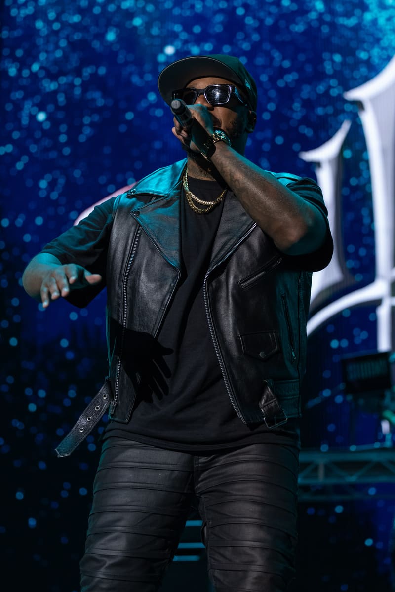 Performer in leather jacket and sunglasses singing into microphone against blue bokeh stage backdrop.