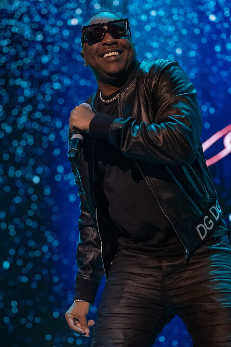 Performer in sunglasses and black leather jacket smiling at microphone against blue bokeh stage backdrop.