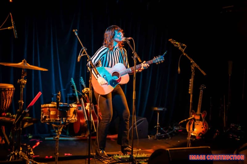 Musician performs with acoustic guitar under warm stage lighting, surrounded by drums and amplifiers on a dark blue curtain backdrop.