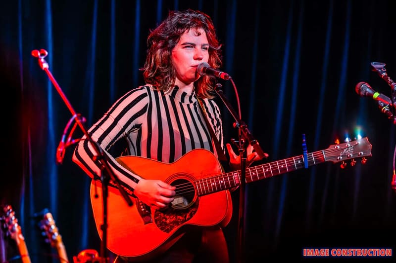 Musician with curly hair playing an acoustic guitar on stage, wearing a striped shirt under warm spotlights.
