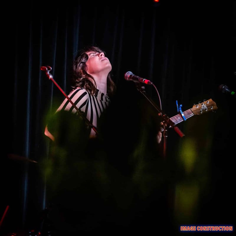 Performer singing at microphone with guitar against dark curtains, lit by stage spotlights.
