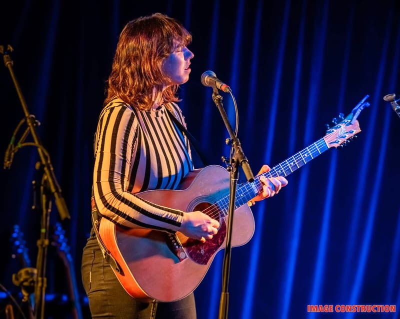 Musician in striped yellow and black shirt playing acoustic guitar at microphone under blue stage lighting.