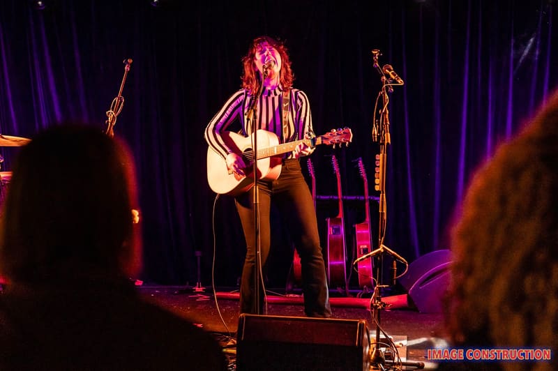 Musician with red hair performing with acoustic guitar under purple stage lighting.