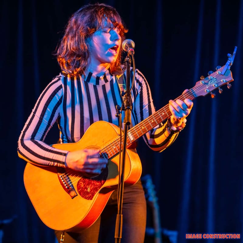 Musician in striped shirt performing with acoustic guitar at microphone under warm and cool stage lighting.