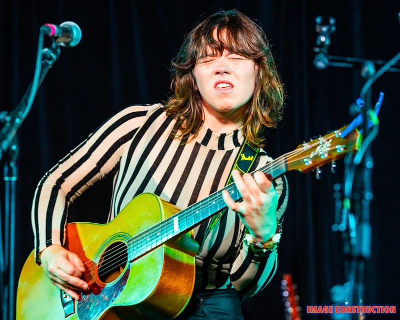 Musician in striped shirt playing acoustic guitar under stage lighting.