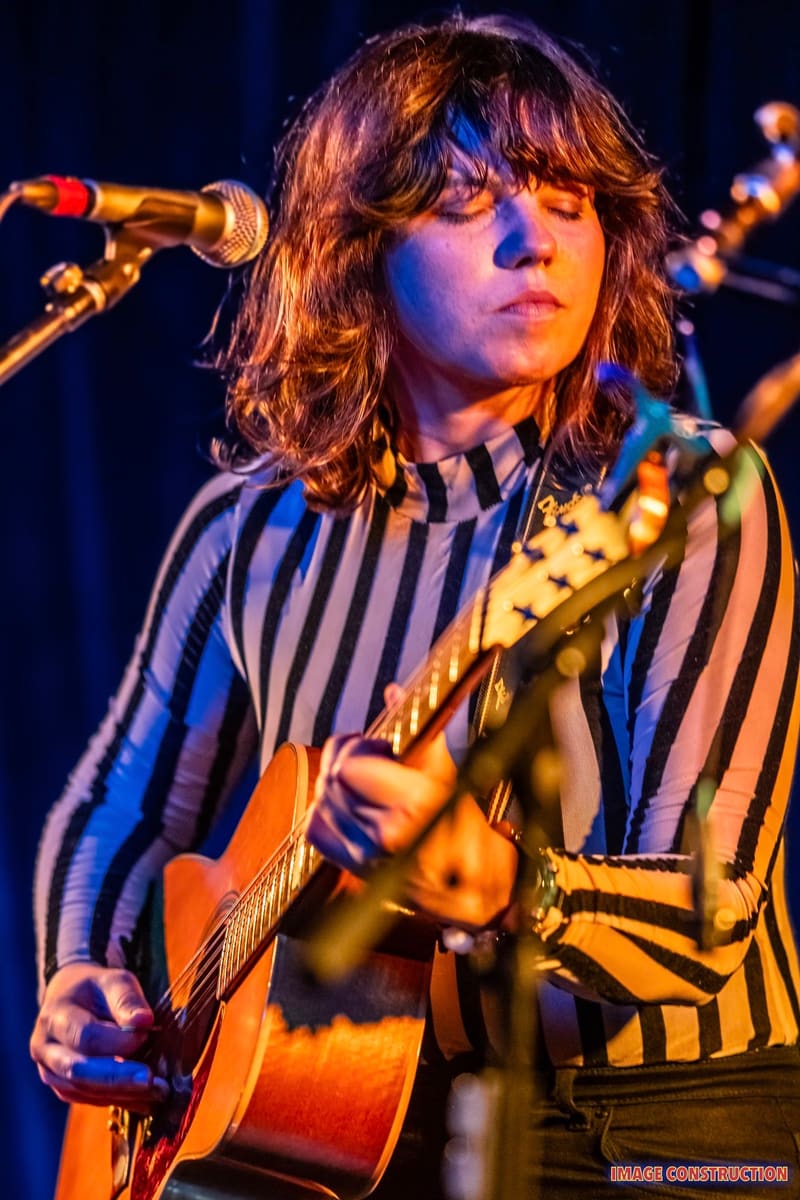 Musician performing with acoustic guitar at microphone, wearing striped shirt under warm and blue stage lighting.