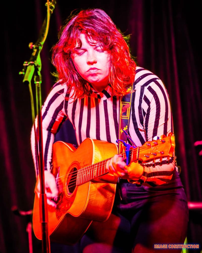 Musician with bright red hair and striped shirt performing with acoustic guitar under warm stage lighting.