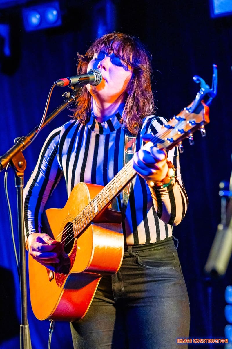 Musician performing at microphone with acoustic guitar, wearing striped shirt under blue stage lighting.