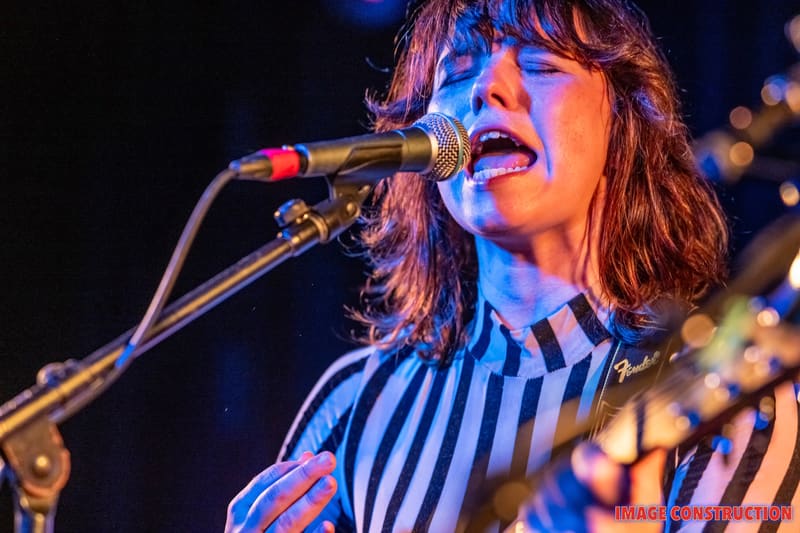 Musician singing into microphone wearing a blue-striped shirt under blue stage lighting.