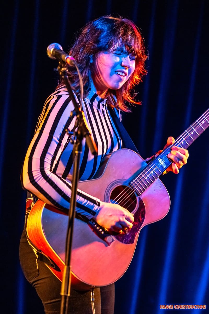 Musician with shaggy dark hair in striped shirt playing acoustic guitar at microphone under blue and red stage lighting.
