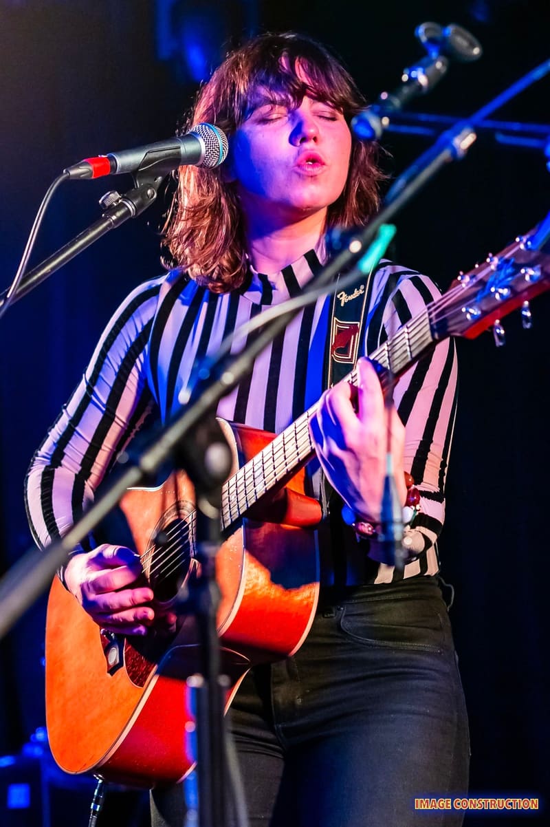 Musician performing at microphone with acoustic guitar, wearing striped shirt under blue stage lighting.