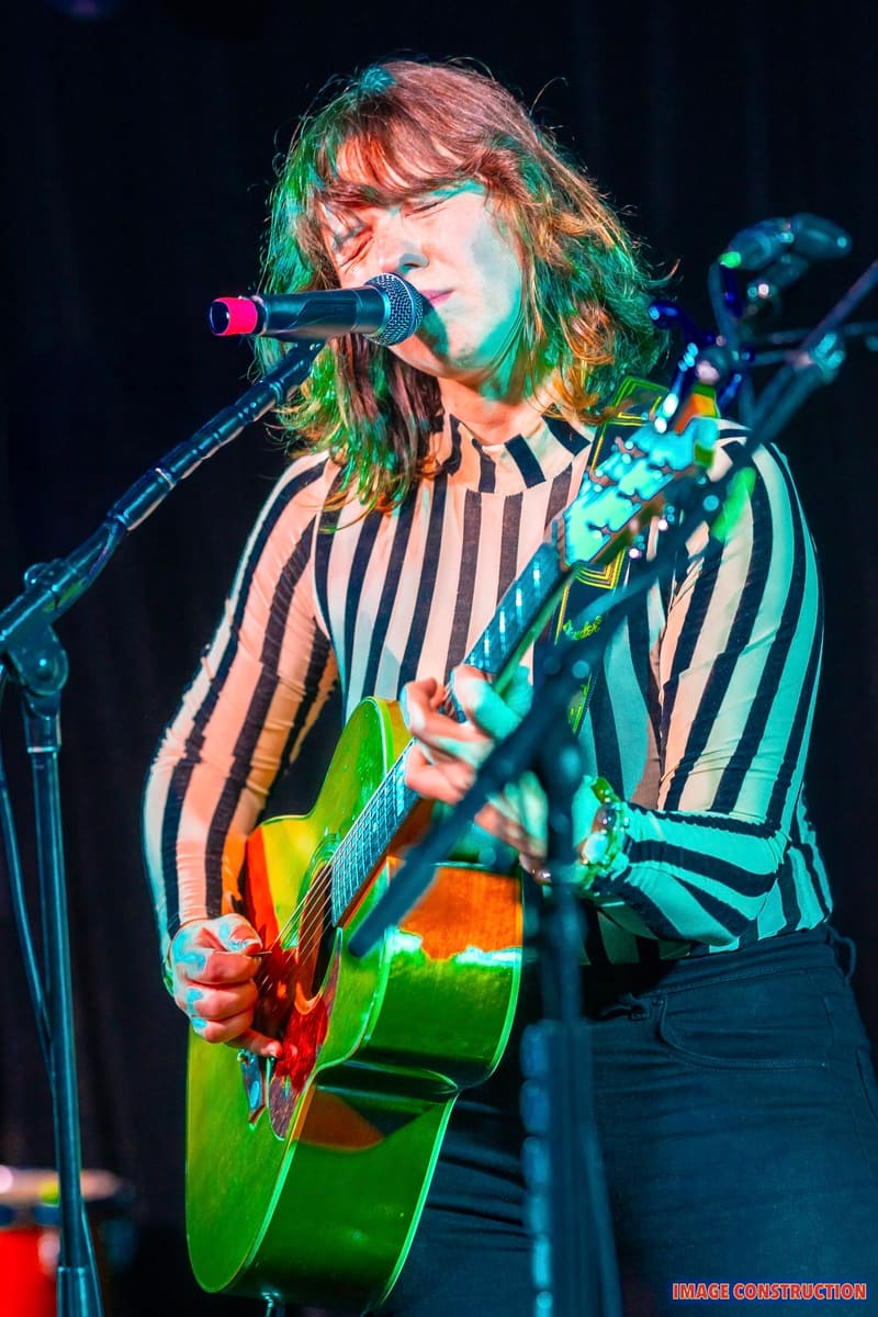 Performer with shoulder-length hair singing into microphone whilst playing acoustic guitar under coloured stage lighting.