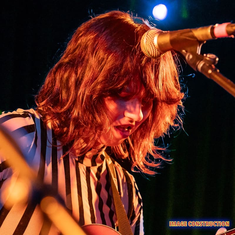 Performer with long reddish hair singing at a microphone under warm stage lighting.