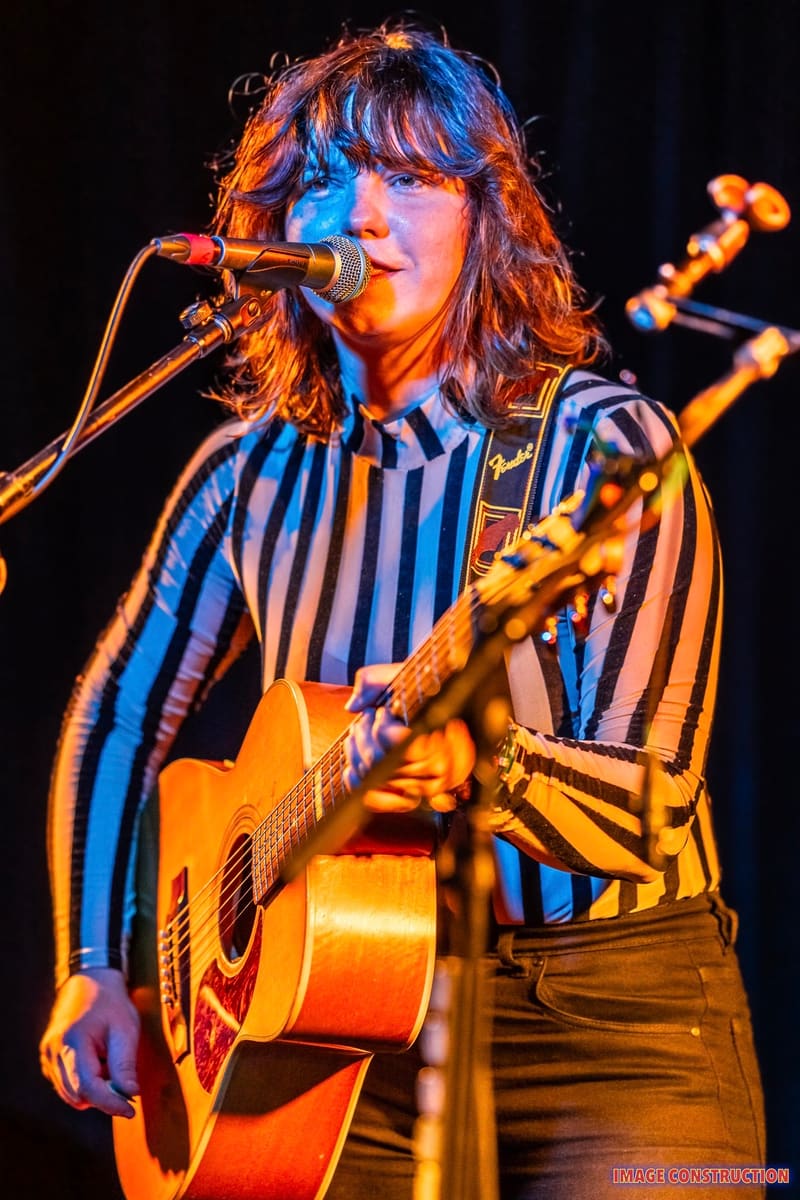 Musician singing into microphone while playing acoustic guitar, lit by warm orange and cool blue stage lighting.