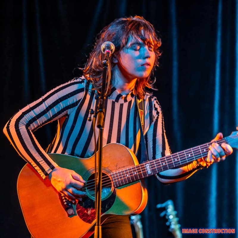 Musician with red hair playing acoustic guitar under warm stage lighting, wearing a striped shirt.
