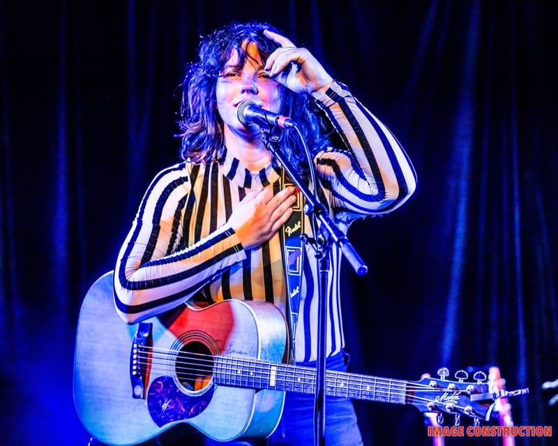 Musician in blue and white striped shirt performing with acoustic guitar under blue stage lighting.