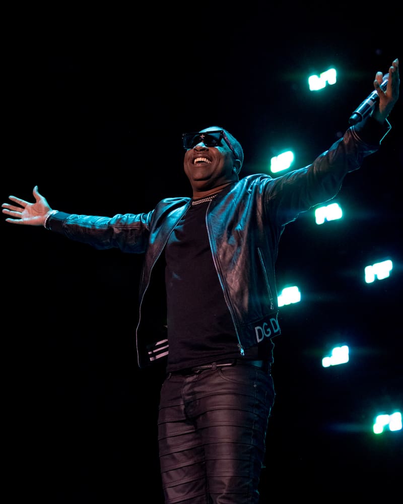 Performer with arms outstretched on stage, lit by turquoise spotlights against a dark background.