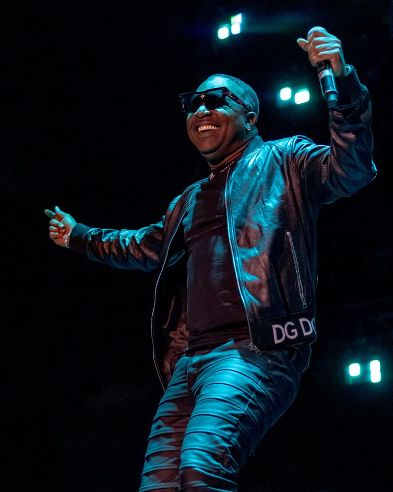 Performer in sunglasses and shiny jacket raises microphone under blue stage lights during live show.