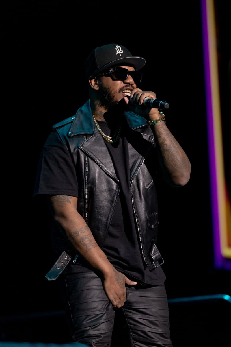 Hip-hop performer in black cap and leather vest singing into microphone under purple stage lighting.