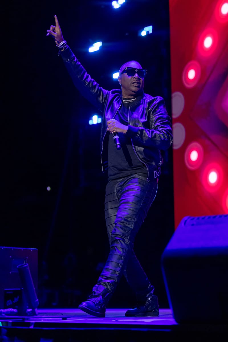 Performer in sunglasses and leather jacket raises fist on stage bathed in blue and red stage lighting.