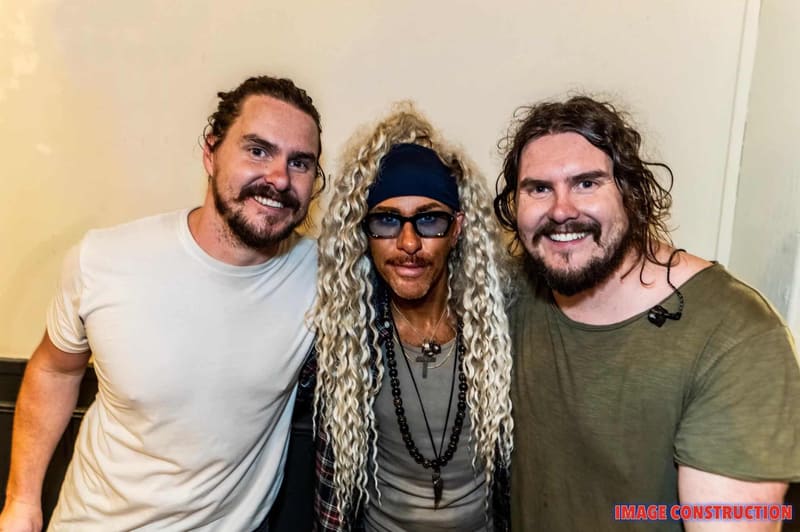Three band members pose together indoors, with the centre figure wearing long blonde braids and sunglasses.