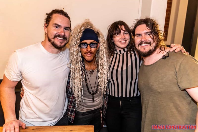 Four people pose together at an indoor event, smiling at the camera with their arms around each other.