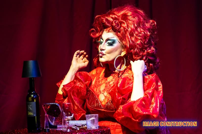 Performer in bright red wig and orange outfit posed at microphone against deep red stage curtain.