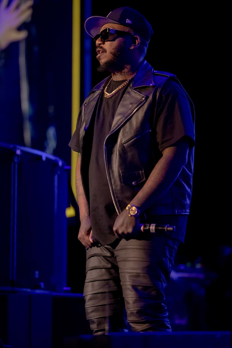 Performer wearing cap and sunglasses holds microphone on stage, bathed in blue lighting.