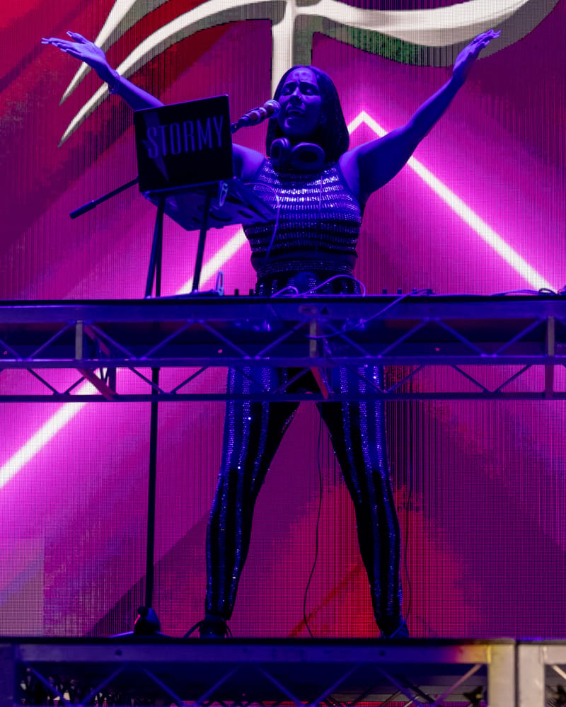 Performer on stage with arms raised, silhouetted against vibrant pink and blue neon lighting.
