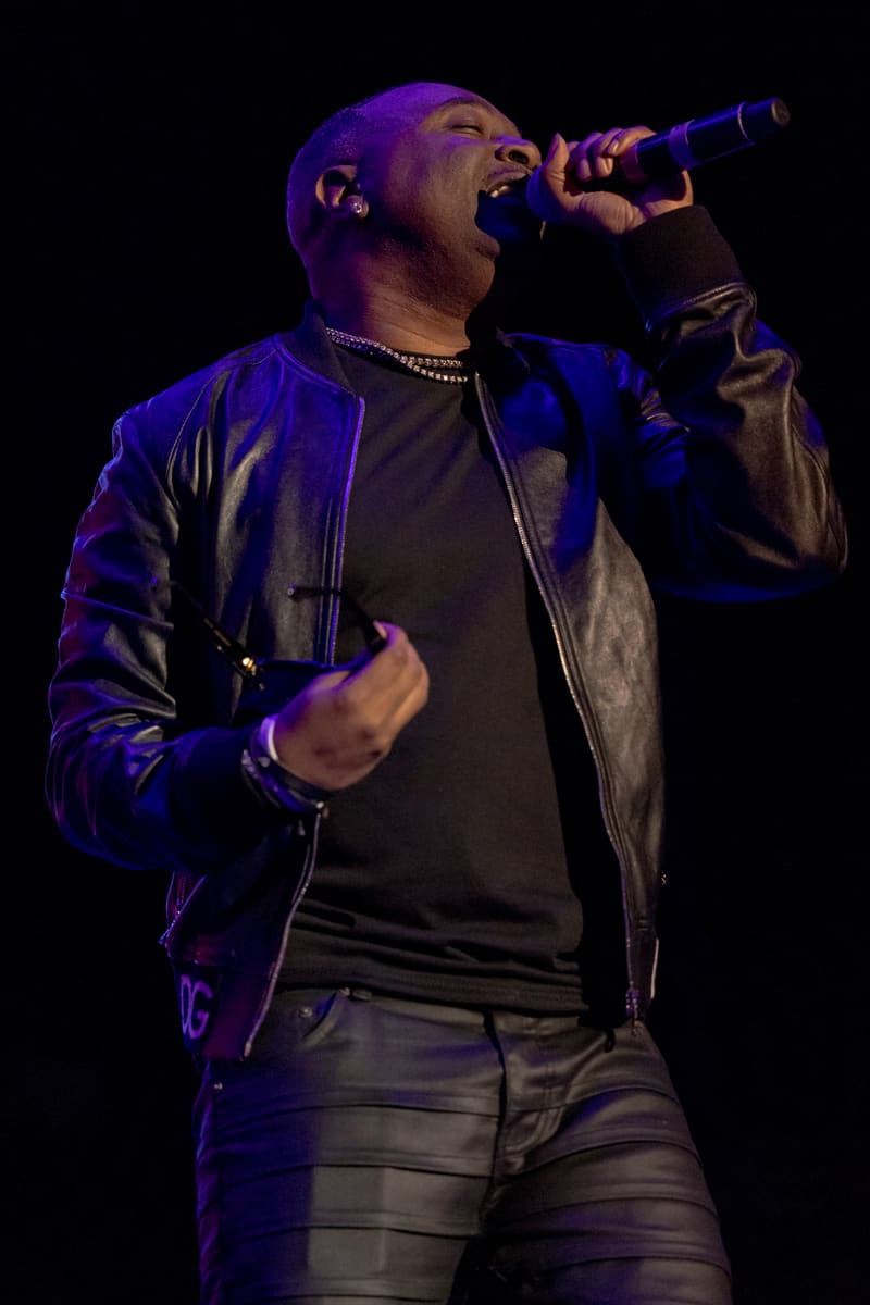 Performer singing into microphone on stage, lit by purple spotlights, wearing leather jacket and chain necklace.