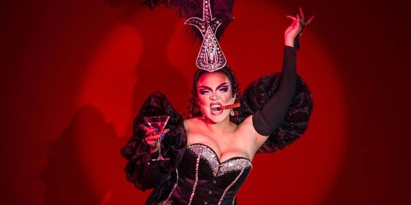 Performer in elaborate black and red costume with ornate headpiece strikes a dramatic pose against a glowing red background.